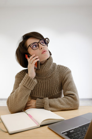 Woman on phone at desk with notebook and laptop in thoughtful poseの写真素材