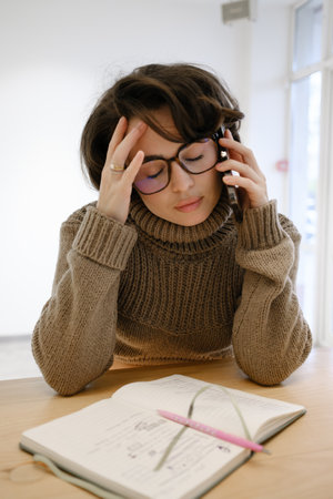 Woman in glasses working at a desk while talking on the phoneの写真素材