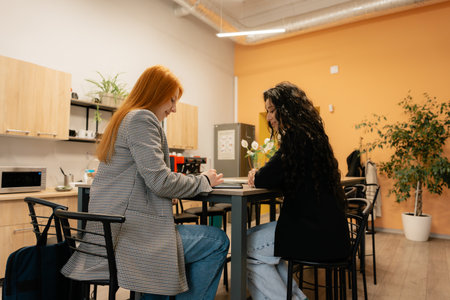 Two women discuss documents at a modern office break area, collaborating on a project together