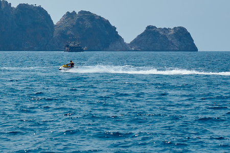 Men in lifejacket riding a white and yellow water scooter on the blue sea, forming large waves, splashes and foam with rocks and ship on the background.の写真素材