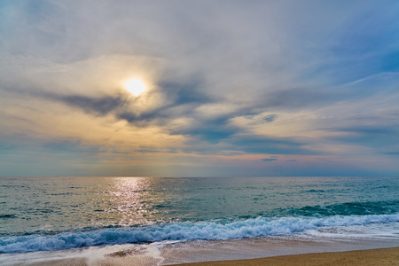 Sunset at the tropical beach, sun behind clouds reflects on water and waves with foam hitting sand.の写真素材