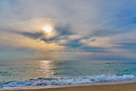 Sunset at the tropical beach, sun behind clouds reflects on water and waves with foam hitting sand.の写真素材