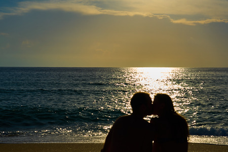 Silhouettes of a couple, man and woman sitting on a sandy beach and kissing with sunset on the sea on a background. Copy space.の写真素材