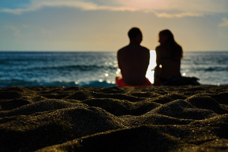 Silhouettes of a couple, man and woman are sitting on a sandy beach and looking at the sunset on the...