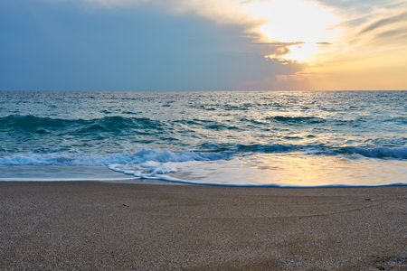 Sunset at the tropical beach, sun behind clouds reflects on water and waves with foam hitting sand.の写真素材