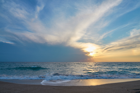 Sunset at the tropical beach, sun behind clouds reflects on water and waves with foam hitting sand.の写真素材