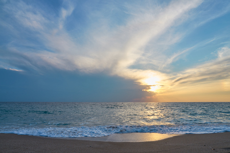Sunset at the tropical beach, sun behind clouds reflects on water and waves with foam hitting sand.の写真素材