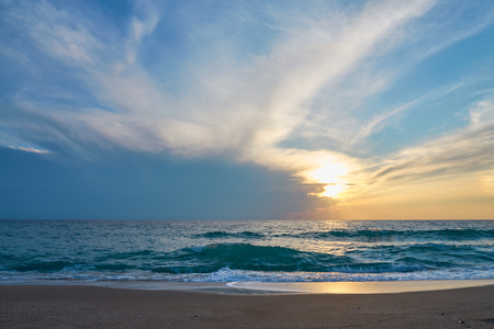Sunset at the tropical beach, sun behind clouds reflects on water and waves with foam hitting sand.の写真素材