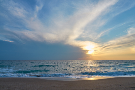 Sunset at the tropical beach, sun behind clouds reflects on water and waves with foam hitting sand.の写真素材
