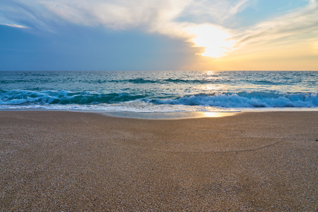 Sunset at the tropical beach, sun behind clouds reflects on water and waves with foam hitting sand.の写真素材