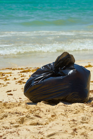 Black plastic garbage bag on a sandy beach with a tropical sea in the background.の写真素材