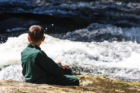 A boy in green clothes watches the flowing waters of a river sitting on a rock. Located in Batistella Reserve: Corupa, Santa Catarina State, Brazil.のeditorial素材