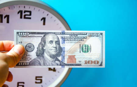 Analog white clock and dollars on a blue background. A man’s hand holds notes of 100 dollars on a...