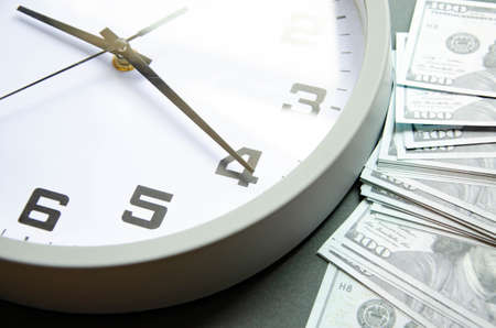 Analog white clock and dollars on a gray, yellow, blue background. Banknotes next to the clock, on...