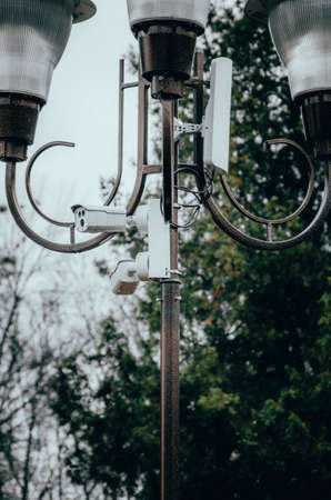 White surveillance camera hanging on a pole. The camera watches the order in the streets and in the parks. Camera on a post in a park. Side view and straightの写真素材