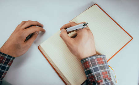 Men's hands, forearm hold a pen, notebook, smartphone. Hands write in notebook in a cell ideas, plans, tasks. Kick on a notebook, break, bend. CopySpace Copy Space Orange, diary. Work with tasks.の写真素材
