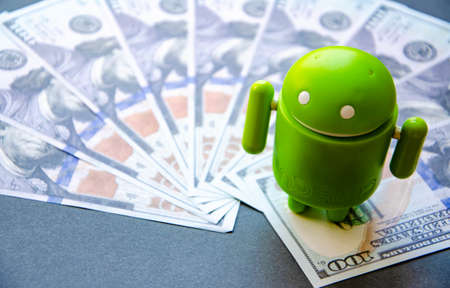 March 2020. Kramatorsk, Ukraine. Green android figurine on different backgrounds, along with dollars. Banknotes dollars in the background. Android and dollarsのeditorial素材