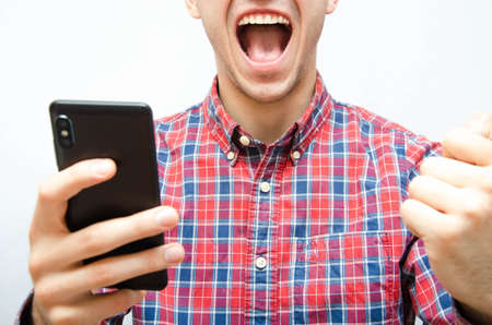 Young man in a plaid shirt with a smartphone on a white background. The guy holds the smartphone in front of him and screams, clenching his fist in front of him with joy. Man rejoices, screaming, winの写真素材