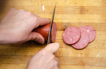 A female hand holds a knife and she cuts sausage on a wooden board. Pizza sausageの写真素材
