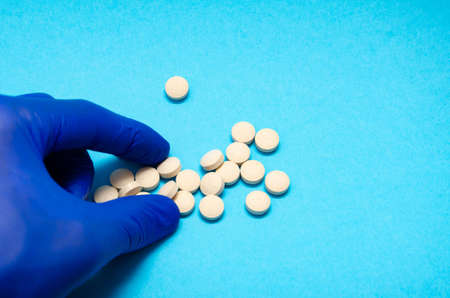 A manâs hand in blue medical gloves takes a tablet with his fingers, on a blue background in close-up. Hand and pillsの写真素材