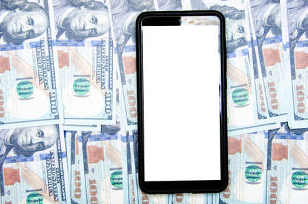 Dollars and smartphone. 100 dollar bills lie throughout the frame on them lies a black smartphone with a white screen on top and at an angleの写真素材