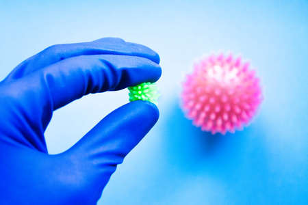 A male hand in blue medical sterile gloves holds a vaccine against coronavirus which is in the background on a blue, yellow, pink background. Anti-influenza agent. Big and small virus. Covid-19の写真素材