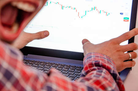 Trading on the exchange. Men's hands in a plaid shirt behind a laptop clenched into a fist. On-screen rates, promotions. Win, lose. Good luck and failure betting, trading. Focus on laptop and graphicsの写真素材