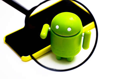 A green android figure on a white background and next to a yellow smartphone a view of it through a magnifying glass. Android and magnifying glass. July 2020. Kiev, Ukraineのeditorial素材