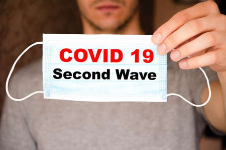 Covid 19 second wave. Covid 19. A man holds a medical mask in front of him on a blue, yellow, black background and the inscription Covid 19 second waveの写真素材