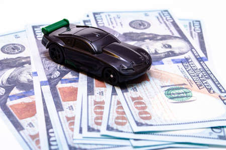 Black toy car on a heap of dollar bills on a white and black background. A sports car with a green spoiler, a wing on dollars in a close-up top and side view. Rear and front car. Selling, buyingの写真素材