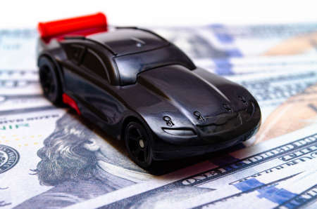 Black toy car on a heap of dollar bills on a white and black background. A sports car with a red spoiler, a wing on dollars in a close-up top and side view. Rear and front car. Selling, buyingの写真素材