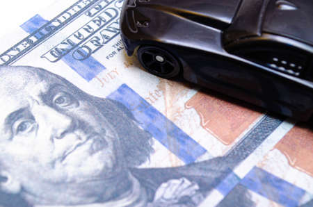 Black toy car on a heap of dollar bills on a white and black background. A sports car with a green spoiler, a wing on dollars in a close-up top and side view. Rear and front car. Selling, buyingの写真素材