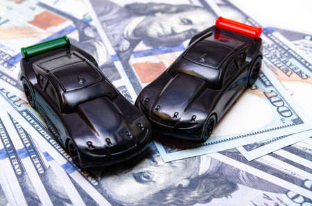 Two black toy cars on a heap of dollar bills on a white and black background. Two sports cars on dollars in close-up. One car overturned, accident, accident. Car insuranceの写真素材