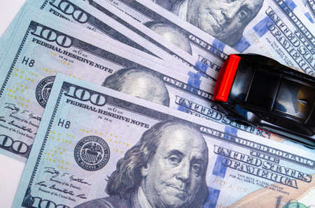 Black toy car on a heap of dollar bills on a white and black background. A sports car with a red spoiler, a wing on dollars in a close-up top and side view. Rear and front car. Selling, buyingの写真素材