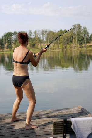 a woman in a bikini throws a fishing pole from a jetty on a lakeの写真素材