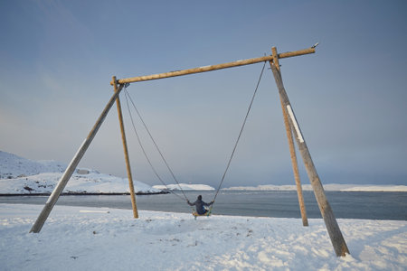 A person in a blue jacket swings on a large swing against the background of the northern sea and snow in winterの写真素材