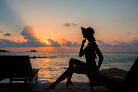 Silhouette of a beautiful slender girl on a sunset background and sun loungers on the ocean shore. Woman watch skyline on sunrise on near deck chair at the sea beach. Vacation Concept.の写真素材