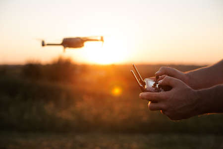 Man holds remote controller with his hands while copter is flying on background. Drone hovers behind the pilot on suset. No faceの写真素材