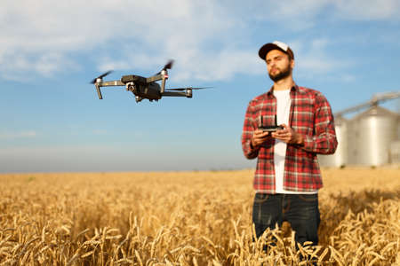 Drone hovers in front of farmer with remote controller in hands near grain elevator. Quadcopter flies near pilot. Agronomist taking aerial photos and videos in a wheat field.の写真素材