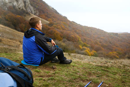 Young tourist hiker with backpack siting and relaxing on the top of the hill in mountains and looking at beautiful yellow autumn landscape sunset sceneryの写真素材