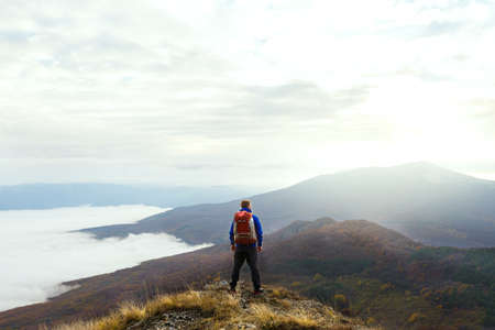 Back view of young tourist hiker with backpack standing on the top of the mountain and looking at beautiful yellow autumn landscape sunset over cloudsの写真素材
