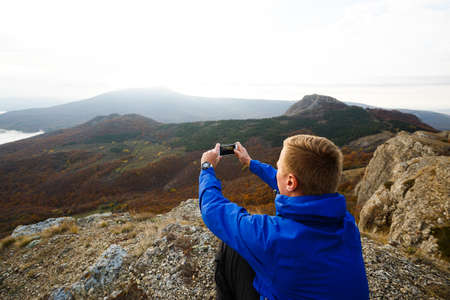 Hiker sitting and taking photo of beautiful mountain landscape with mobile phone. Climber man photographing panorama with smartphone on summit.の写真素材