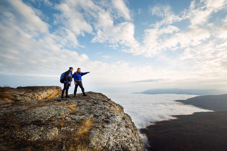 Two climbers standing on top of summit above clouds in the mountains. Hiker man pointing with his...