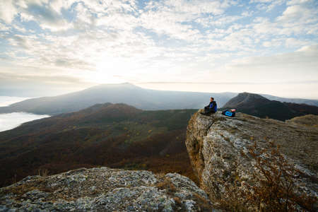 Smiling man hiker with backpack siting and relaxing on the top of the mountain and looking at beautiful yellow autumn landscape sunset over cloudsの写真素材