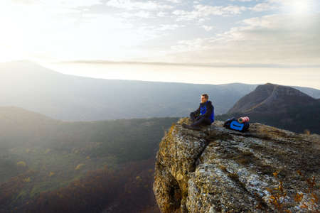 Smiling man hiker with backpack siting and relaxing on the top of the mountain and looking at beautiful yellow autumn landscape sunset over cloudsの写真素材