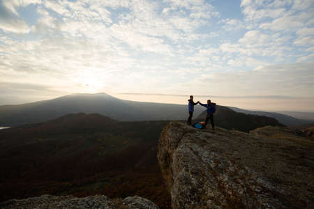 Two climbers standing on summit above clouds in the mountains holding hands. Silhouettes of hikers celebrating ascent on sunset. Help, cooperation, synergy, friendship concept.の写真素材