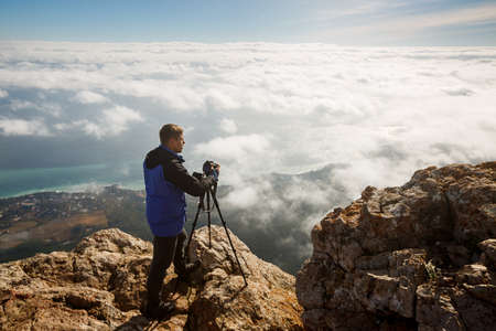 Man standing with a tripod and camera on a high mountain peak above clouds, city and sea....
