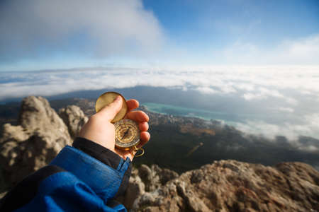 Point of view photo of explorer man searching direction with golden compass in his hand above clouds with autumn mountains and sea backgroundの写真素材