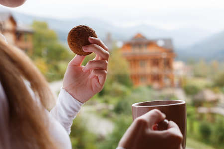 Woman holds cup of coffee and oat cookie in her hands at mountain resort. Female with tea mug and snack with forest and wooden hotel on background. Dietary concept.の写真素材