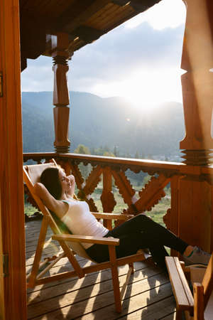 Woman is lying on wooden deck chair and relaxes looking at beautiful mountain resort scenic on sunset. Young slim female on terrace wooden balcony sitting on chaise-longue at cozy country houseの写真素材